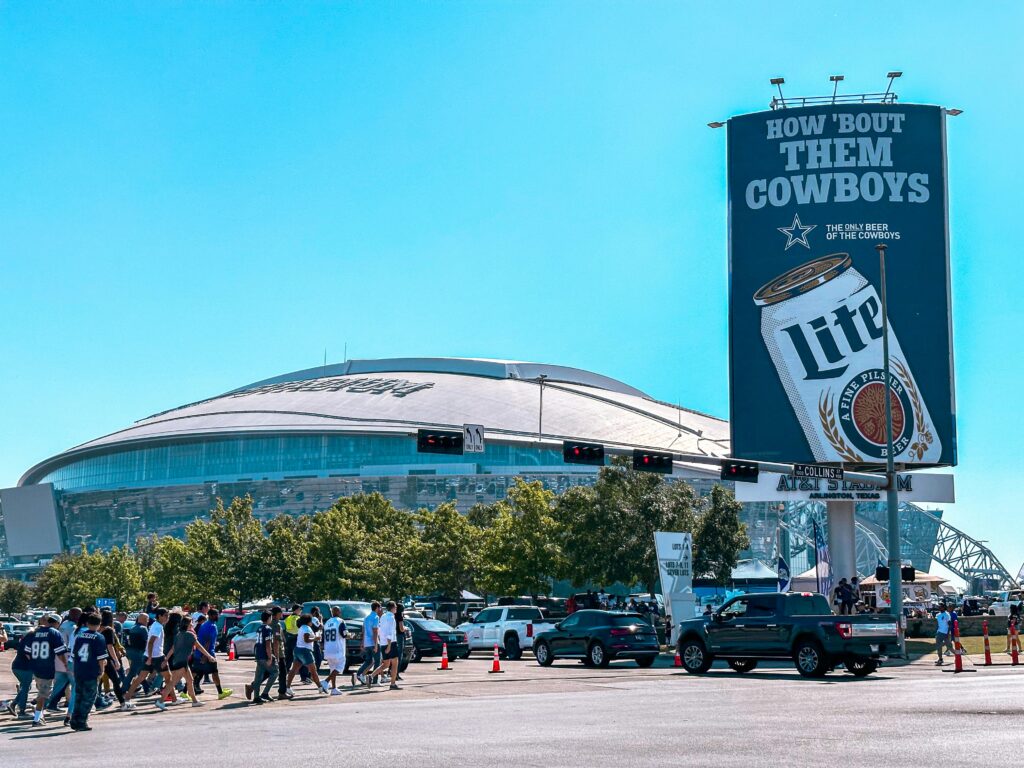 People walking near AT&T Stadium on a sunny day in Arlington, Texas, with a prominent billboard.